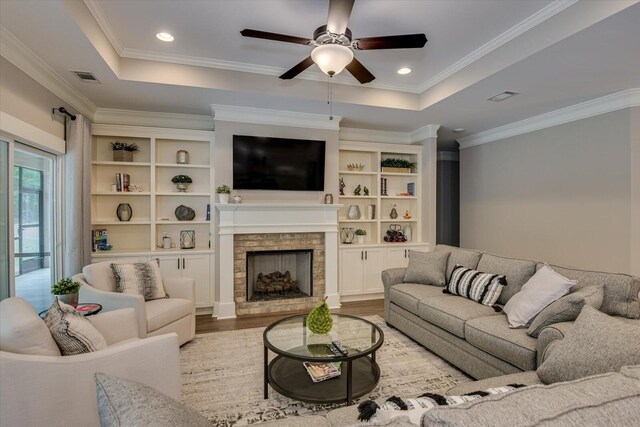 living room featuring a raised ceiling, ceiling fan, crown molding, hardwood / wood-style floors, and a stone fireplace