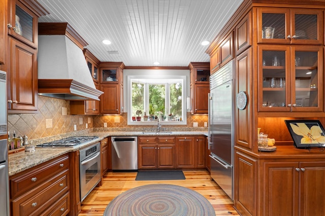 kitchen with appliances with stainless steel finishes, custom exhaust hood, light wood finished floors, light stone countertops, and brown cabinetry
