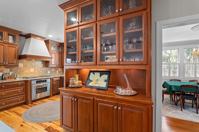 kitchen with appliances with stainless steel finishes, light wood-style flooring, custom exhaust hood, tasteful backsplash, and brown cabinets