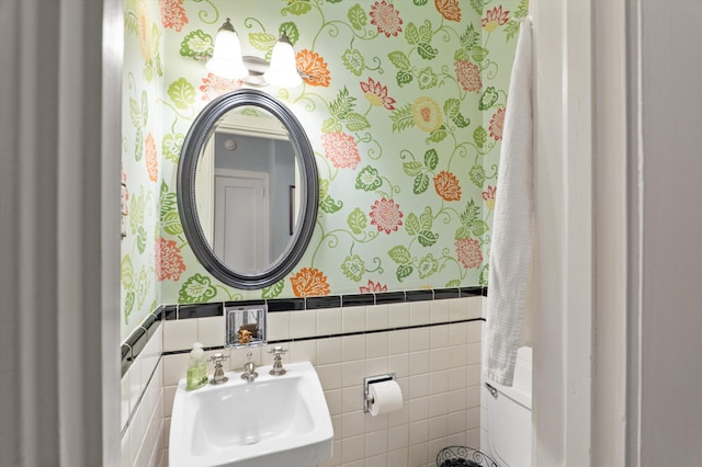 half bath with a sink, wallpapered walls, wainscoting, and tile walls