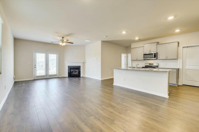 kitchen featuring a kitchen island with sink, ceiling fan, light stone countertops, appliances with stainless steel finishes, and wood-type flooring