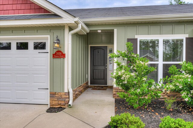 view of exterior entry featuring an attached garage, board and batten siding, roof with shingles, and stone siding