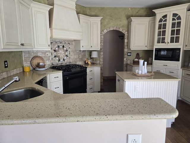 kitchen with light stone counters, premium range hood, a sink, black appliances, and dark wood-type flooring