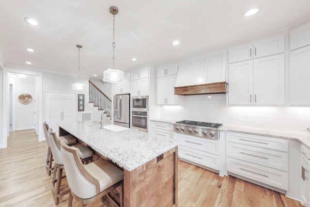 kitchen with white cabinetry, decorative light fixtures, a center island with sink, appliances with stainless steel finishes, and light wood-type flooring