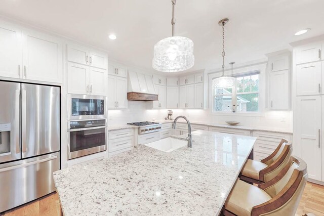 kitchen with sink, stainless steel appliances, an island with sink, decorative light fixtures, and custom range hood