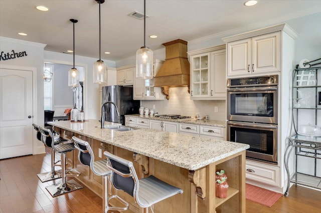 kitchen featuring premium range hood, hanging light fixtures, an island with sink, a breakfast bar area, and stainless steel appliances