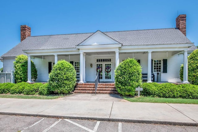 greek revival inspired property featuring covered porch, french doors, roof with shingles, uncovered parking, and a chimney