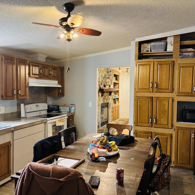 dining area featuring crown molding, ceiling fan, a textured ceiling, light tile patterned flooring, and a stone fireplace