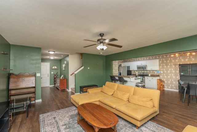 living room featuring ceiling fan, baseboards, wallpapered walls, dark wood-style floors, and stairway