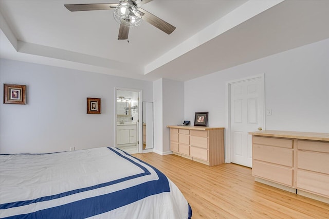 bedroom with a tray ceiling, ensuite bath, light wood-style flooring, and a ceiling fan