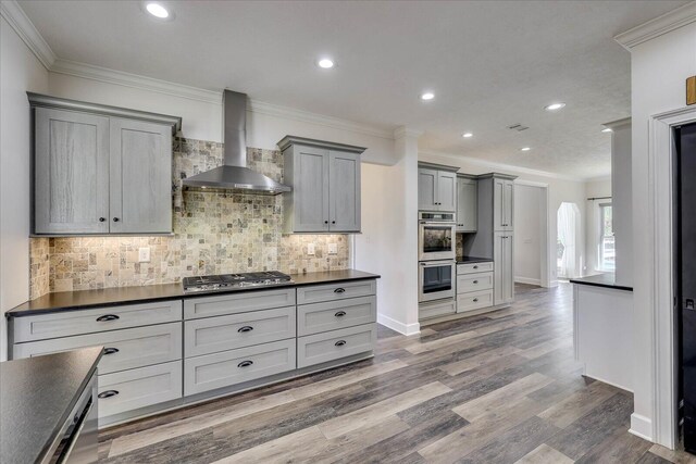 kitchen with wall chimney exhaust hood, gray cabinetry, appliances with stainless steel finishes, ornamental molding, and hardwood / wood-style flooring