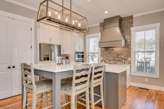 kitchen with white cabinets, ornamental molding, custom exhaust hood, hanging light fixtures, and appliances with stainless steel finishes