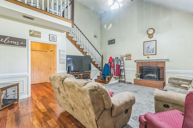 living room featuring a brick fireplace, wood finished floors, high vaulted ceiling, stairs, and a wainscoted wall