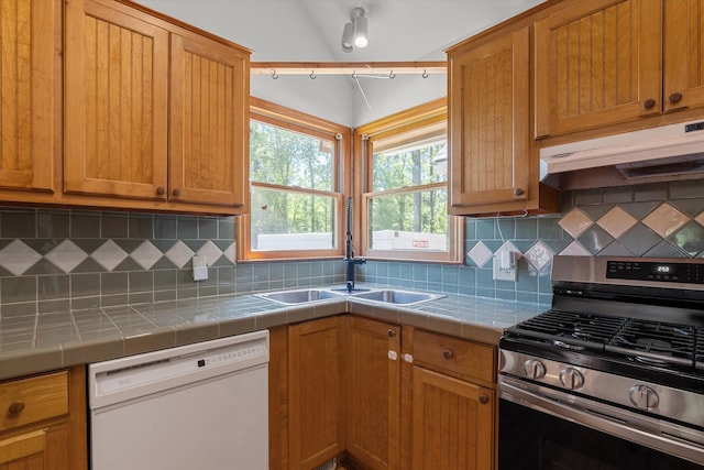 kitchen with a sink, white dishwasher, under cabinet range hood, tile countertops, and stainless steel gas range oven