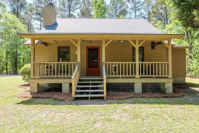 view of front facade with a chimney, a front yard, a shingled roof, and covered porch