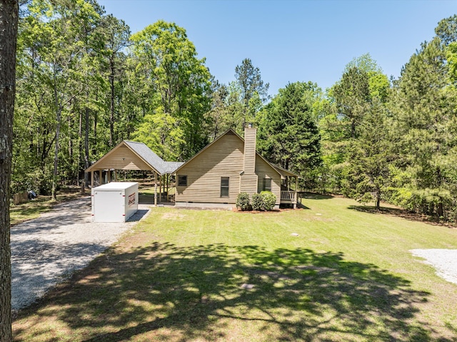 view of front facade featuring a chimney, a carport, a view of trees, a front yard, and gravel driveway