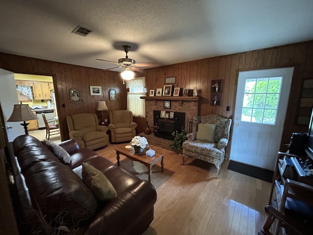living room with wooden walls, ceiling fan, wood-type flooring, a textured ceiling, and a brick fireplace