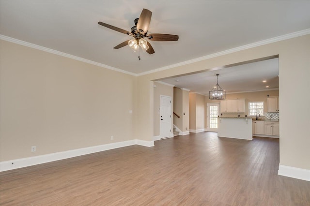 unfurnished living room with baseboards, wood finished floors, crown molding, a sink, and ceiling fan with notable chandelier