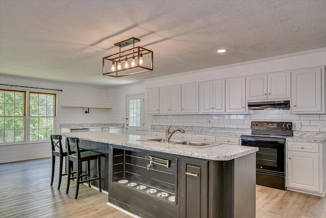 kitchen with black range with electric cooktop, under cabinet range hood, a sink, open shelves, and a center island with sink