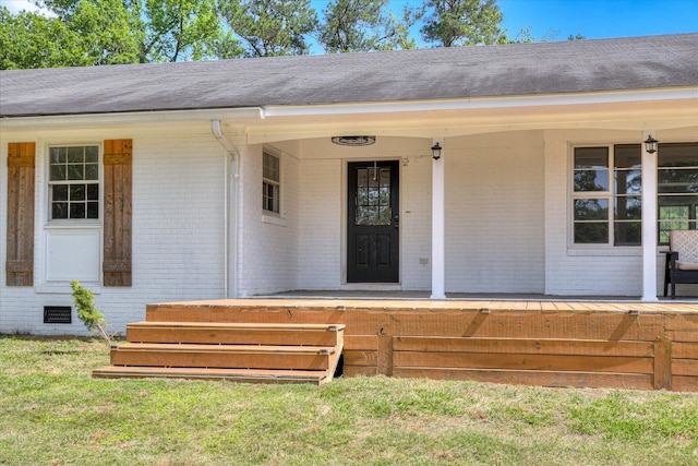 doorway to property featuring brick siding, covered porch, roof with shingles, and a lawn