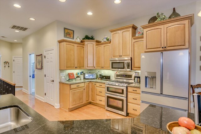 kitchen featuring dark stone countertops, light brown cabinetry, light hardwood / wood-style flooring, and appliances with stainless steel finishes