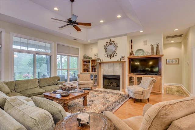 living room with a tray ceiling, a tiled fireplace, ceiling fan, and light wood-type flooring