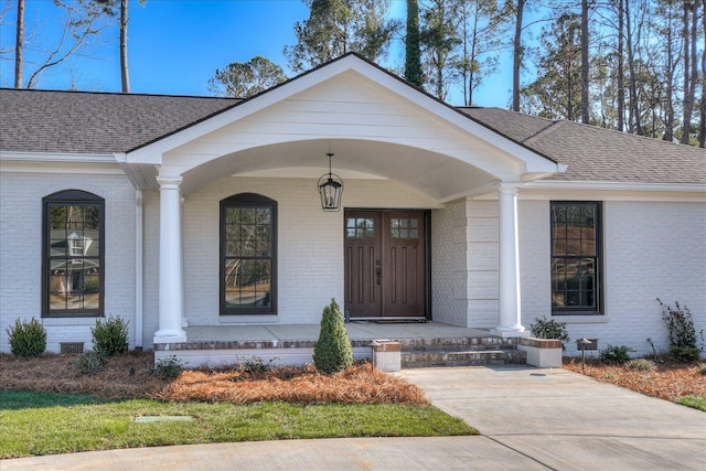 view of exterior entry with crawl space, covered porch, a shingled roof, and brick siding