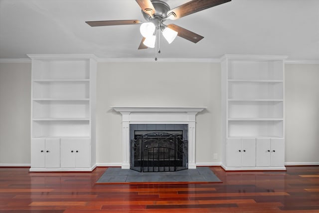 unfurnished living room featuring a tile fireplace, built in features, ceiling fan, crown molding, and dark wood-type flooring