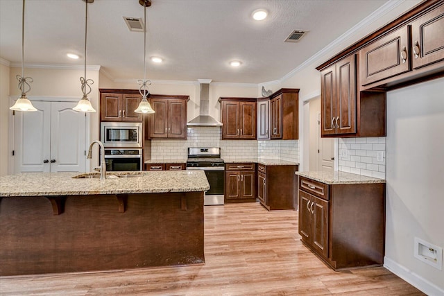 kitchen with wall chimney exhaust hood, stainless steel appliances, decorative light fixtures, and a kitchen island with sink