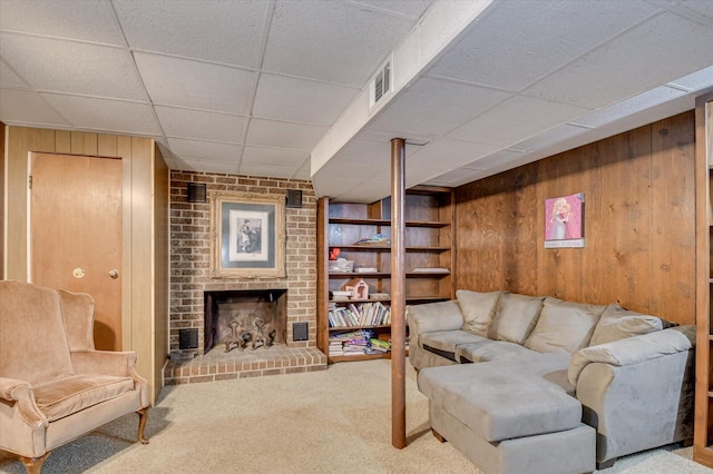 carpeted living room with a paneled ceiling, wooden walls, and a brick fireplace