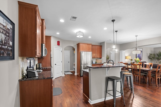 kitchen featuring appliances with stainless steel finishes, arched walkways, brown cabinets, a kitchen bar, and dark wood-style floors