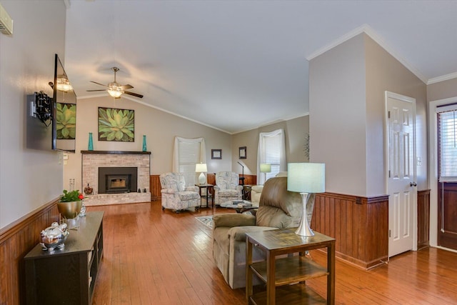 living room with ceiling fan, vaulted ceiling, a brick fireplace, light wood-type flooring, and ornamental molding