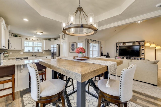 dining room featuring sink, a chandelier, vaulted ceiling, a tray ceiling, and light wood-type flooring