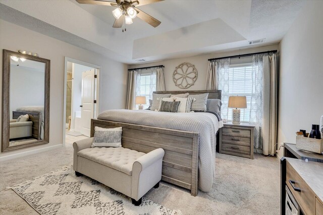 bedroom with ceiling fan, ensuite bathroom, light carpet, and a tray ceiling