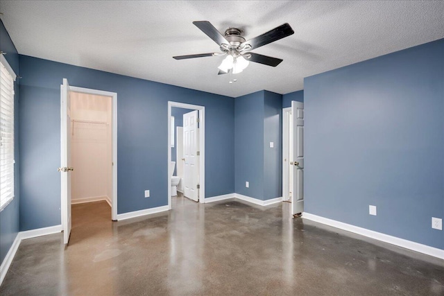 unfurnished bedroom with finished concrete flooring, baseboards, a walk in closet, ceiling fan, and ensuite bath
