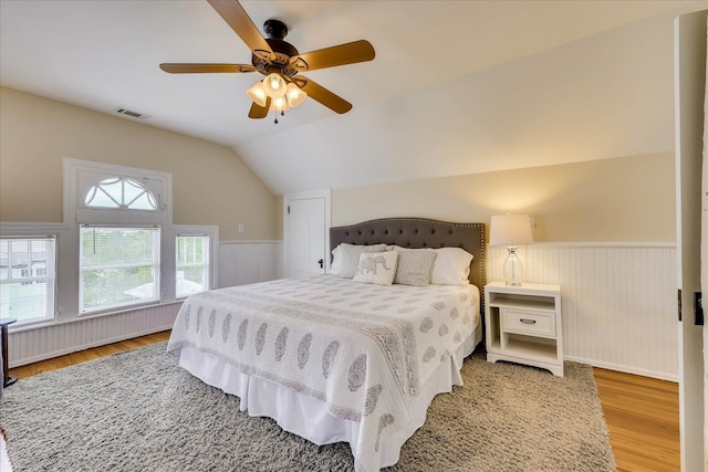 bedroom with light wood-type flooring, a ceiling fan, a wainscoted wall, visible vents, and lofted ceiling