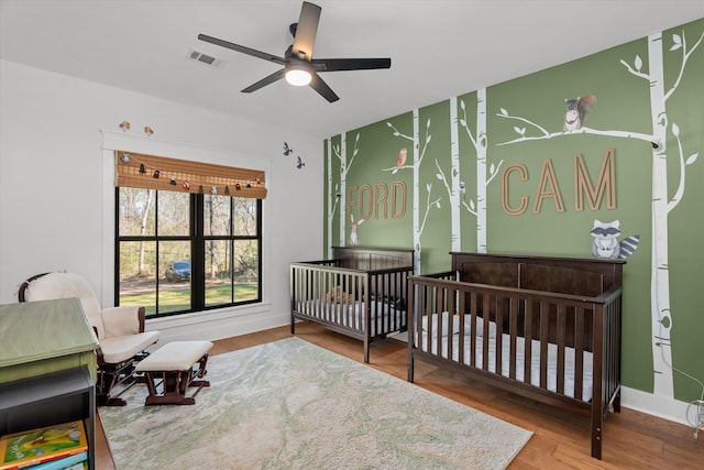 bedroom with wood finished floors, visible vents, baseboards, ceiling fan, and a nursery area