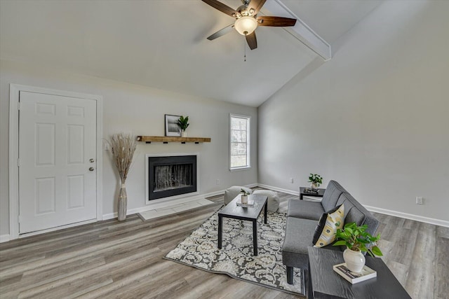 living area featuring a ceiling fan, wood finished floors, baseboards, lofted ceiling with beams, and a fireplace with flush hearth