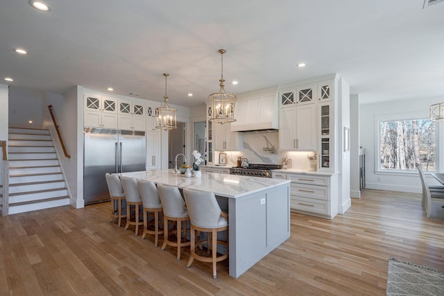 kitchen with a kitchen island with sink, stainless steel built in refrigerator, white cabinets, decorative light fixtures, and custom exhaust hood