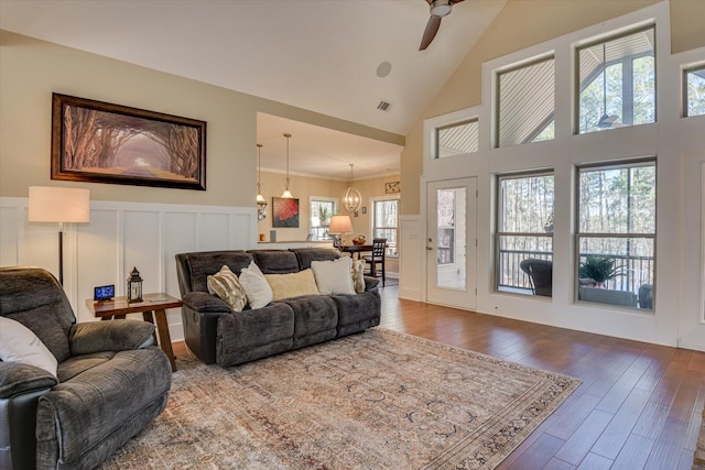 living area featuring visible vents, high vaulted ceiling, ceiling fan with notable chandelier, wood finished floors, and a decorative wall