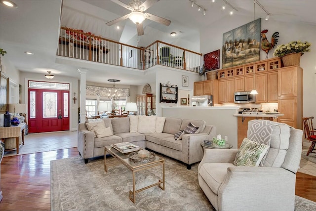 living room with ceiling fan with notable chandelier, rail lighting, wood finished floors, and high vaulted ceiling