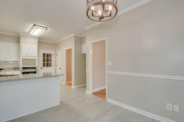 kitchen with light stone counters, ornamental molding, a notable chandelier, white cabinets, and oven