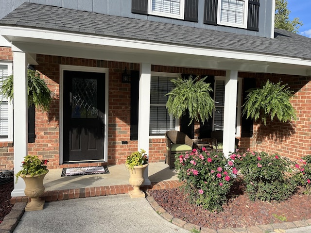 entrance to property featuring brick siding, covered porch, and roof with shingles