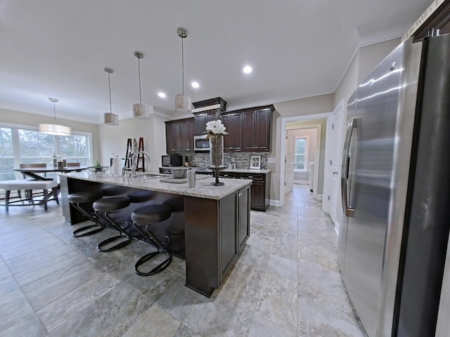 kitchen with light stone countertops, hanging light fixtures, tasteful backsplash, a center island with sink, and appliances with stainless steel finishes