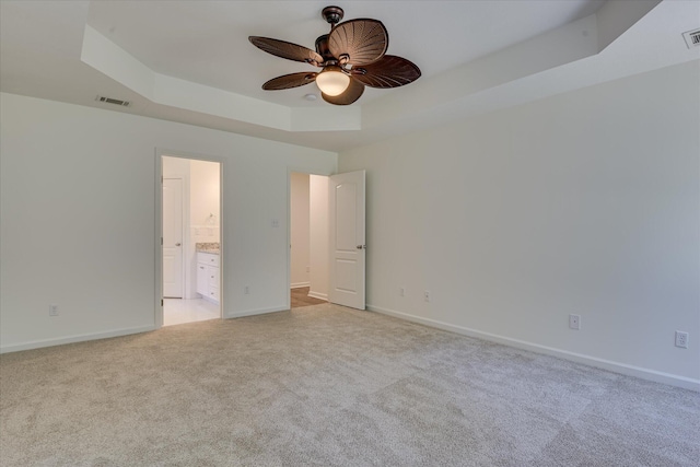 unfurnished bedroom with a tray ceiling, light carpet, baseboards, connected bathroom, and ceiling fan