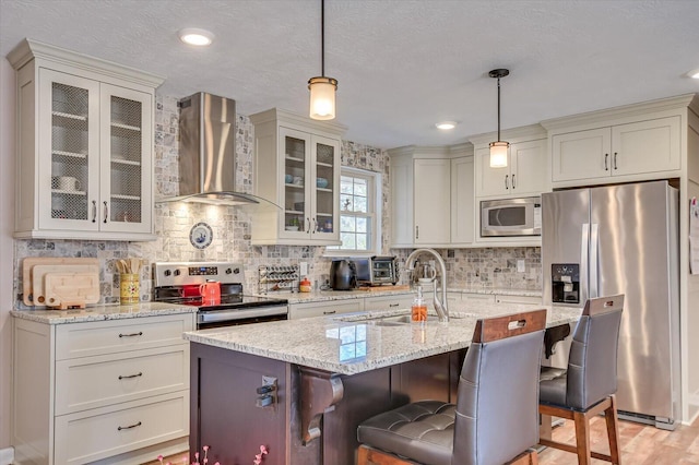 kitchen featuring hanging light fixtures, appliances with stainless steel finishes, wall chimney exhaust hood, and a kitchen island with sink