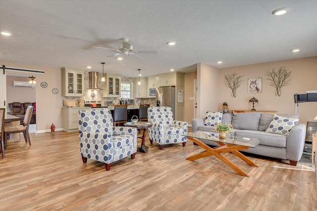 living room with a textured ceiling, light wood-type flooring, ceiling fan, a barn door, and a wall mounted air conditioner