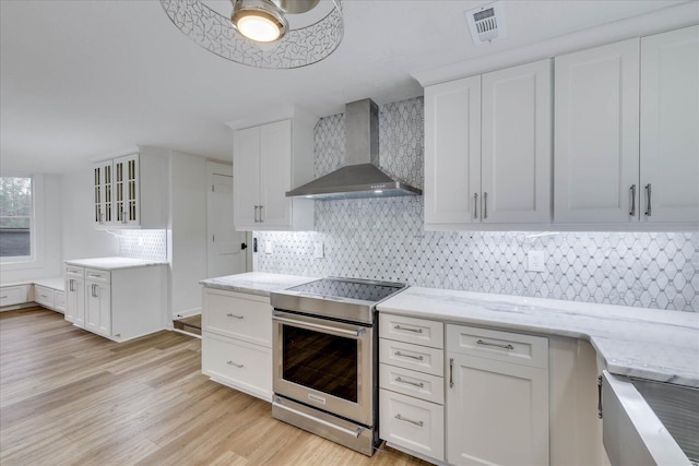 kitchen featuring wall chimney exhaust hood, light hardwood / wood-style floors, stainless steel electric range, and white cabinets