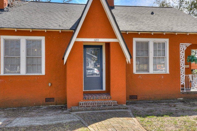 property entrance with crawl space, stucco siding, visible vents, and roof with shingles