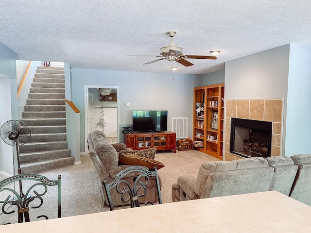 carpeted living room with a textured ceiling, ceiling fan, visible vents, a tiled fireplace, and stairs